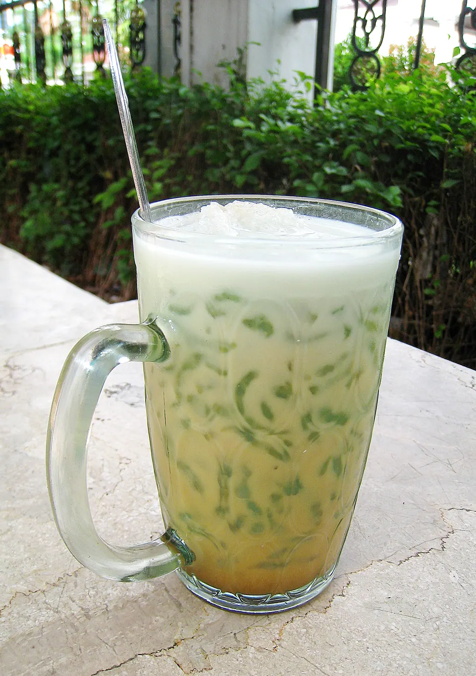 Segelas es cendol: untaian cendol hijau dalam santan, dengan langit dan dedaunan di belakangnya